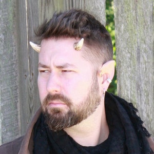 May include: A man with short brown hair wearing a brown jacket and a black scarf. He has a beard and is wearing small horns on his forehead. He is also wearing a silver hoop earring in his left ear.