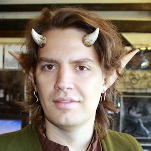 May include: A person with brown hair wearing a brown shirt with a green jacket and a necklace with a green stone pendant. The person is wearing fake elf ears and horns on their forehead.