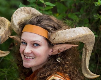 May include: A person wearing a costume with large, curved horns, a tan headband, and pointed ears. The costume includes a brown and tan striped vest, a rust-colored scarf, and a lace sleeve. The person is smiling, with curly brown hair.