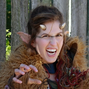 May include: A woman with pointed ears and horns is wearing a brown fur coat with a skull necklace. She is making a claw-like gesture with her hand.