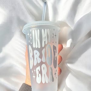 May include: A clear plastic tumbler with a lid and straw. The tumbler is held by a person and features the text "IN MY BRIDE ERA" in a wavy, silver font, with diamond ring graphics. The theme is bridal.