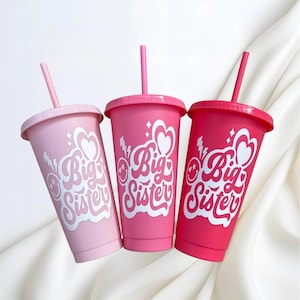 May include: Three pink tumblers with lids and straws, each with the text "Big Sister" in a white, retro font. The tumblers are in shades of light pink, medium pink, and hot pink.