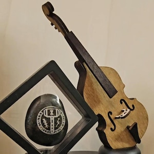 May include: A black stone with a silver engraved design of a hammer and lightning bolt inside a circle with runes is displayed in a black diamond-shaped display case. A wooden miniature violin with a black neck is displayed on a black stand next to the display case.