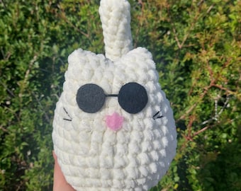May include: A white, plush cat toy with black sunglasses and a pink heart-shaped mouth. The cat has black stitched whiskers and a tall, upright tail. The toy is made of a soft, textured material and is held against a backdrop of green foliage.