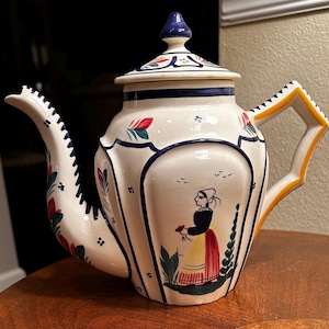 May include: A white ceramic teapot with a blue and yellow handle and lid. The teapot is decorated with a colorful floral pattern and a woman in a red dress holding flowers.