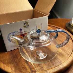 May include: A clear glass teapot with a blue handle and lid, sitting on a wooden surface. The teapot is in front of a cardboard box with the word "TEAVANA" printed on it. The teapot has a capacity of approximately 1 liter.