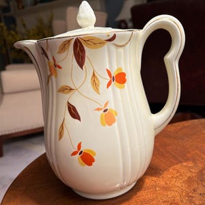 May include: A white ceramic teapot with a brown and gold floral pattern. The teapot has a lid with a small knob on top. The teapot is sitting on a wooden table.
