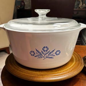 May include: White casserole dish with a blue floral design and a clear glass lid. The dish is sitting on a wooden surface.