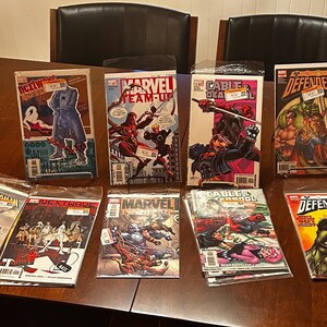 May include: A collection of comic books featuring various Marvel characters, including Spider-Man, Cable, and the Defenders. The comics are in protective sleeves and displayed on a wooden table. Some comics are propped up on stands.