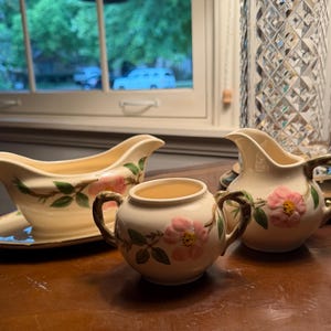 May include: A cream-colored ceramic gravy boat with an attached underplate, a sugar bowl with two handles, and a creamer pitcher. Each piece features a hand-painted design of pink flowers with yellow centers and green leaves on brown stems.