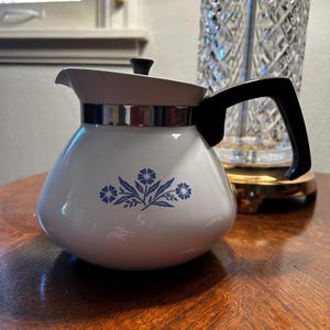 May include: White enamel teapot with a black handle and a silver band around the top. The teapot has a blue and white floral design on the front.
