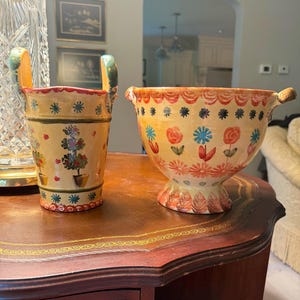 May include: A tall, cylindrical ceramic pitcher with two handles and a shorter, wide-mouthed ceramic bowl, both featuring a cream-colored base with hand-painted floral motifs in red, blue, and green, and scalloped decorative trim along the edges.