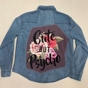 May include: A light blue denim long-sleeve shirt featuring a large, dark purple fabric patch on the back. The patch displays a floral arrangement of pink and red roses with green leaves and the black text "Cute BUT Psycho" printed over them.