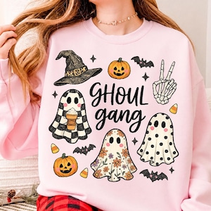 Peut inclure: Un sweat-shirt rose clair avec une impression graphique portant le texte "GHOUL gang" en écriture noire. Le motif comprend un chapeau de sorcière, deux citrouilles d'Halloween, trois fantômes à motifs, une main squelette faisant un signe de paix, des chauves-souris et des bonbons en forme de maïs.
