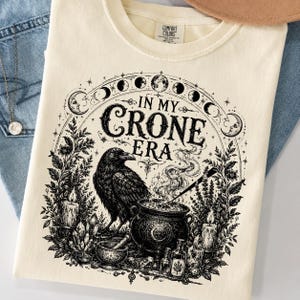 May include: Cream-colored t-shirt with a black graphic featuring a raven, cauldron, and the text "IN MY CRONE ERA." The design includes phases of the moon, candles, and botanical elements, creating a mystical theme.