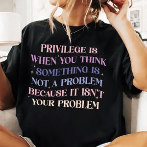 May include: Black t-shirt with the text "PRIVILEGE IS WHEN YOU THINK SOMETHING IS NOT A PROBLEM BECAUSE IT ISN'T YOUR PROBLEM" in pink, purple, and blue lettering.