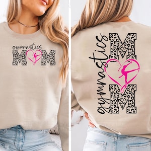 Peut inclure: Un sweat-shirt beige avec l'inscription "gymnastics mom" imprimée sur le devant. Les lettres sont noires et soulignées d'un motif léopard. Un cœur rose avec une silhouette de gymnaste est au centre des lettres. Le dos du sweat-shirt porte l'inscription "gymnastics" imprimée verticalement en noir.