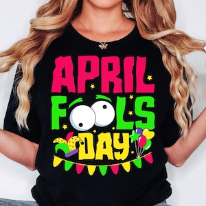 Puede incluir: Camiseta negra con un gráfico colorido que dice "April Fools Day" con un gorro de bufón, ojos saltones y globos.