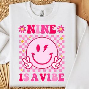 Peut inclure: Sweat-shirt blanc avec un fond à carreaux rose et blanc. Le texte "NINE IS A VIBE" est écrit en lettres roses. Un smiley rose avec un éclair est au centre du motif.