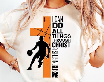 May include: Cream-colored t-shirt featuring a basketball-themed design. An orange cross and basketball graphic are combined with a silhouette of a basketball player. The text reads "I CAN DO ALL THINGS THROUGH CHRIST WHO STRENGTHS ME."