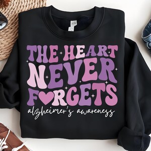 May include: Black sweatshirt with a purple, pink, and white graphic that says "The Heart Never Forgets" with a heart symbol and the text "Alzheimer's Awareness" underneath.