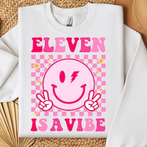 Op de afbeelding: Witte sweatshirt met de tekst "ELEVEN IS A VIBE" in roze. Het ontwerp toont een roze smiley met een bliksemschicht en vredestekens, tegen een roze en witte geruite achtergrond met gele sterren.