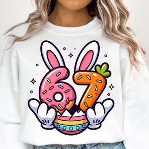 Peut inclure: Sweat-shirt blanc avec un graphisme coloré sur le thème de Pâques. Le motif présente les chiffres "6" et "7" stylisés en beignet et carotte, respectivement, avec des oreilles de lapin et un œuf de Pâques. Le motif est souligné en noir avec des accents roses, oranges et bleus.
