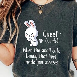 May include: Dark gray t-shirt featuring a white cartoon bunny with the text "Queef: (verb) When the small cute bunny that lives inside you sneezes."