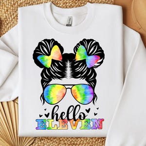 Peut inclure: Sweat-shirt blanc avec un motif représentant une coiffure de dessin animé avec des nœuds et des lunettes de soleil arc-en-ciel. Le texte "hello ELEVEN" est imprimé en couleurs arc-en-ciel en dessous. Le motif est centré sur le devant.