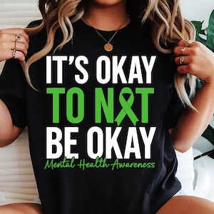 May include: Black t-shirt with white text that reads "It's okay to not be okay" and a green ribbon. The text "Mental Health Awareness" is written below the ribbon.