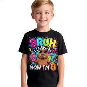Puede incluir: Una camiseta negra con un gráfico de un capibara marrón con gafas de sol, sosteniendo un dónut rosa con forma de número 6 y un dónut azul con forma de número 7. El texto dice BRUH I SURVIVED 6 7 NOW I'M 8 en letras de colores brillantes.