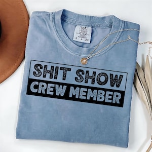 Könnte beinhalten: Ein hellblaues T-Shirt mit dem Text "SHIT SHOW CREW MEMBER" in schwarzen Buchstaben. Der Text hat einen abgenutzten, Vintage-Look.