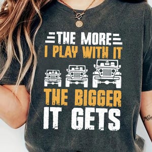 Könnte beinhalten: Dunkelgraues T-Shirt mit dem Text "THE MORE I PLAY WITH IT THE BIGGER IT GETS" in Weiß und Orange. Das Design zeigt drei Jeep-Illustrationen. Ein lässiges, bequemes T-Shirt.