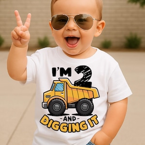 May include: A white t-shirt with a yellow dump truck graphic and the text "I'M 2 AND DIGGING IT". The shirt is worn by a child wearing aviator sunglasses and giving a peace sign.