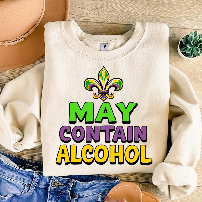May include: A cream-colored sweatshirt with a green, purple, and gold fleur-de-lis design. The text "MAY CONTAIN ALCOHOL" is printed in green, purple, and yellow letters.