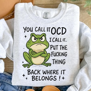 Peut inclure: Sweat-shirt gris clair avec un dessin de grenouille verte et le texte "YOU CALL IT OCD I CALL IT.. PUT THE FUCKING THING BACK WHERE IT BELONGS !+". La grenouille a l'air en colère.