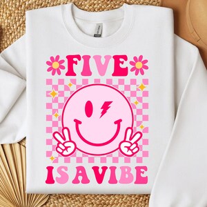 Puede incluir: Sudadera blanca con un diseño a cuadros rosa y blanco. El gráfico incluye el texto "FIVE IS A VIBE" con una carita sonriente, signos de paz y flores.