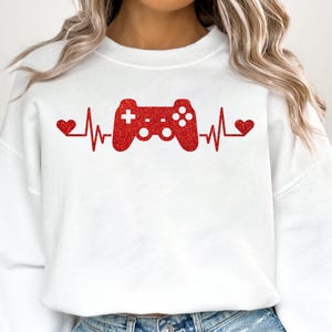 May include: White sweatshirt with a red glitter design of a video game controller and a heartbeat with hearts on either side.