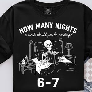 Op de afbeelding: Zwarte sweatshirt met een witte afbeelding van een skelet dat in bed leest, met een nachtkastje, lamp en kaarsen. Tekst luidt "HOW MANY NIGHTS a week should you be reading?" en "6-7".