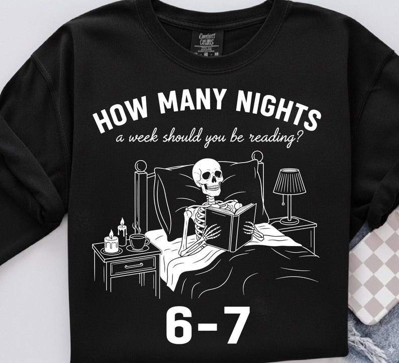 Op de afbeelding: Zwarte sweatshirt met een witte afbeelding van een skelet dat in bed leest, met een nachtkastje, lamp en kaarsen. Tekst luidt "HOW MANY NIGHTS a week should you be reading?" en "6-7".