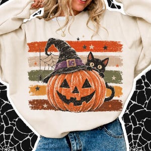 Peut inclure: Un sweat-shirt à col rond couleur crème avec une impression graphique représentant une citrouille orange portant un chapeau de sorcière noir à bande violette. Un chat noir est assis derrière la citrouille, près d'une toile d'araignée.