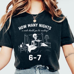 Op de afbeelding: Zwart T-shirt met een witte afbeelding van een skelet dat in bed leest, onder een lamp. De tekst luidt "HOW MANY NIGHTS a week should you be reading?" en "6-7". Een casual kledingstuk.