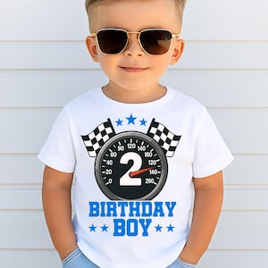 Peut inclure: Un t-shirt blanc avec un motif sur le thème des courses pour un anniversaire de 2 ans. Le motif comprend un compteur de vitesse avec le chiffre "2", des drapeaux à damier et le texte "BIRTHDAY BOY" en bleu.