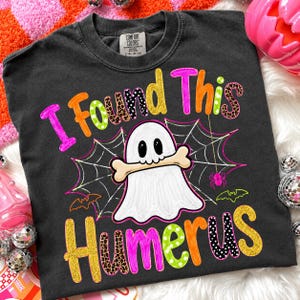 Peut inclure: Un t-shirt gris anthracite avec un motif de fantôme blanc tenant un os dans la bouche. Le texte I FOUND THIS HUMERUS est écrit en lettres colorées et à motifs. Une toile d'araignée, une petite araignée rose et deux chauves-souris sont imprimées.