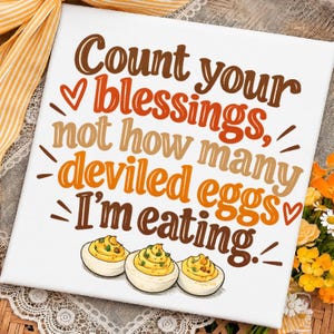 Peut inclure: Un panneau carré blanc avec le texte brun et orange "Count your blessings, not how many deviled eggs I'm eating." Le design comprend de petits cœurs et une illustration de trois œufs farcis en bas du panneau.