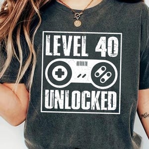 May include: Dark gray t-shirt with a white graphic that reads "LEVEL 40 UNLOCKED" above a controller design. The shirt is a casual style, suitable for everyday wear.