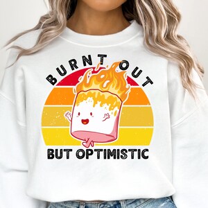 May include: White sweatshirt with a graphic of a marshmallow on fire, with the words "BURNT OUT" above and "BUT OPTIMISTIC" below. The marshmallow has a smiling face and is set against a sunset background.