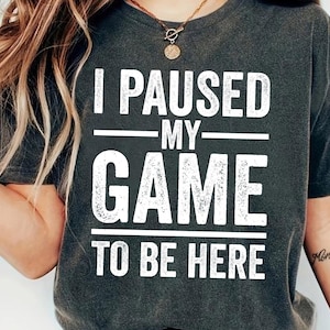 May include: Dark gray t-shirt with the white text "I PAUSED MY GAME TO BE HERE". The shirt has short sleeves and a crew neck. The text is in a bold, distressed font.