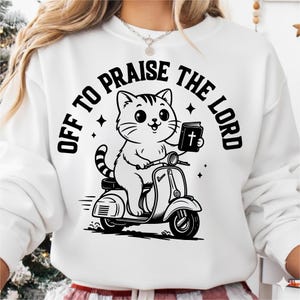 Peut inclure: Sweat-shirt blanc avec un motif noir et blanc d'un chat sur un scooter tenant un livre avec une croix. Le texte "OFF TO PRAISE THE LORD" est en arc au-dessus du chat.