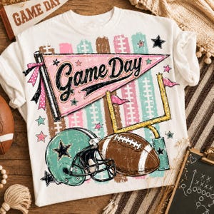Peut inclure: Un t-shirt blanc avec une impression graphique représentant un fanion rose portant le texte Game Day, un casque de football américain turquoise, un ballon de football marron et des poteaux de but jaunes, agrémenté d'étoiles et de rubans.